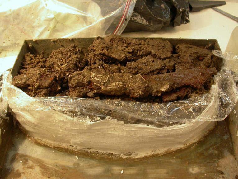 Ett antal fragment av brända ben samt träkolspartiklar var fastkorroderade på svärdet. BROTT Preparat med svärd i fyllfat vid ankomst till konserveringen.