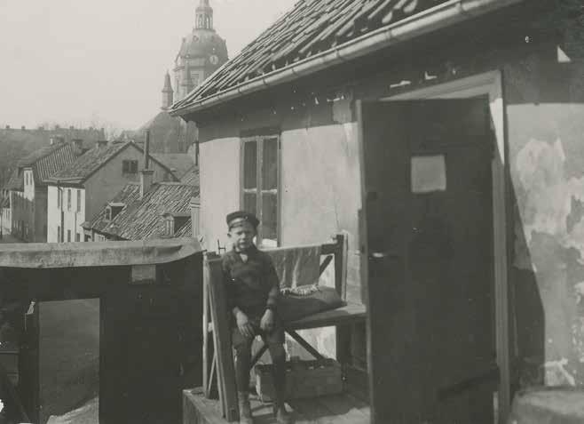 EN POJKE SITTER PÅ BÄNKEN FRAMFÖR STENHUSET OMKRING SEKELSKIFTET 1900. BEBYGGELSEN I BAKGRUNDEN HAR SEDAN DESS ERSATTS AV HÖGRE BYGGNADER. FOTO: O. ÅKERLIND. SSM:S ARKIV, F 66037.