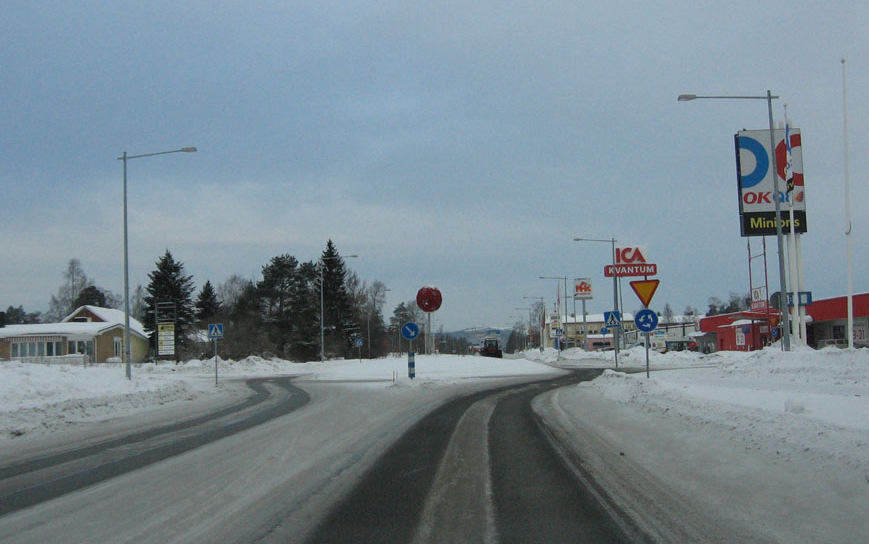 Bebyggelsen blir något tätare med en förstadsmässig prägel. En liten rondell bryter vägens raka sträckning och höga skyltar för OKQ8, ICA, Max och Shell radar upp sig invid vägen.