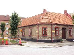 Gulgrått fogstruket tegel. Valmat sadeltak, rött 1-kupigt tegel. Röda 2- och 3-lufts fönster med överbåge.