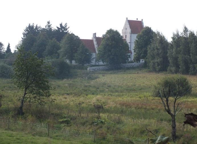 Törringe kyrka från norr med Sege å i förgrunden. Kyrkogårdsmuren består av fogad gråsen, norra sidan. Den östra sidan skyddas av en klippt hagtornshäck.