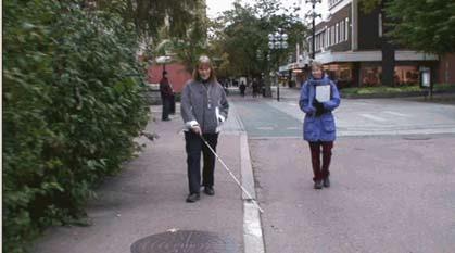 Gör riktningsändring höger 90 grader och följer kant genom att dra käppen längs trottoarkanten över angöringsgata. Stannar till vid två tillfällen innan testpersonen kommer fram till cykelbanan.