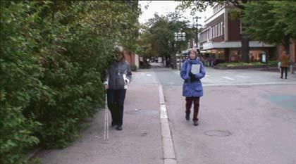 Går inledningsvis på vägen men går sedan upp på trottoaren och följer kantstenen en bit genom att dra käppen längs trottoarkanten och går sedan över till att försöka följa kantstödet som täcks av