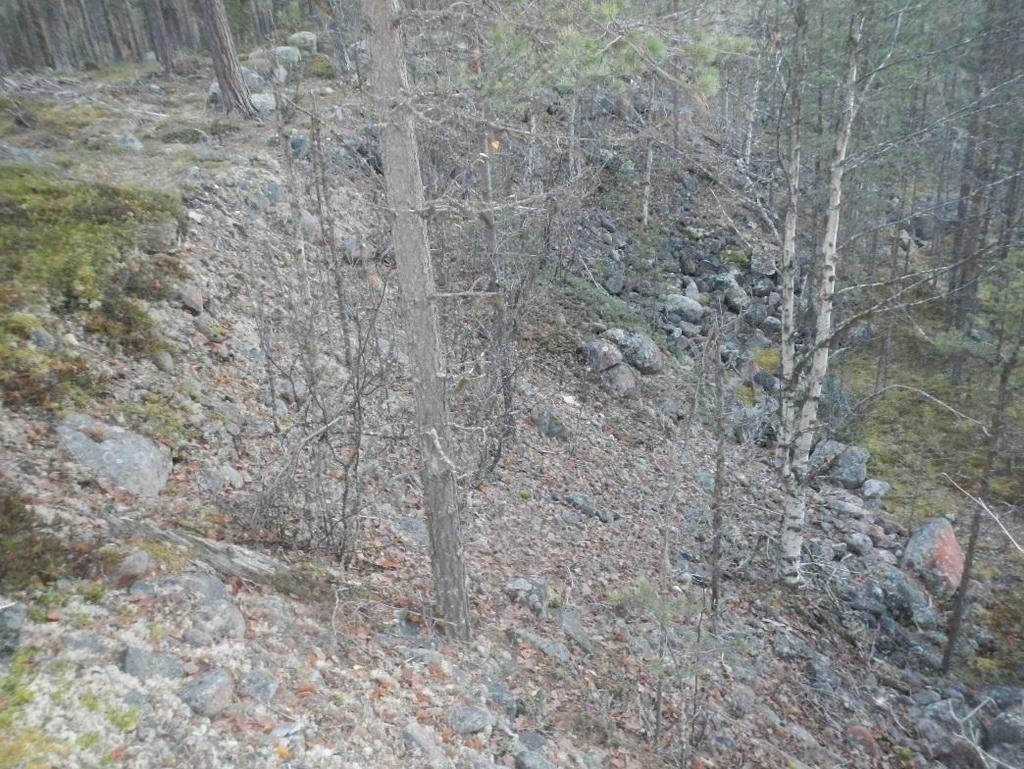 Foto. Slänt ned mot rullskidbanan i sydöst. Grus sten och block kan tydligt urskiljas. Slänten uppskattas vara cirka meter hög.