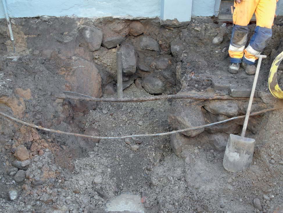 Figur 5. Den påträffade grundmuren till höger i bild, under en äldre elkabel med täckande tegelstenar. I fonden syns Nordénska gårdens grundmur. Foto mot öster av Mats Magnusson, ÖM.