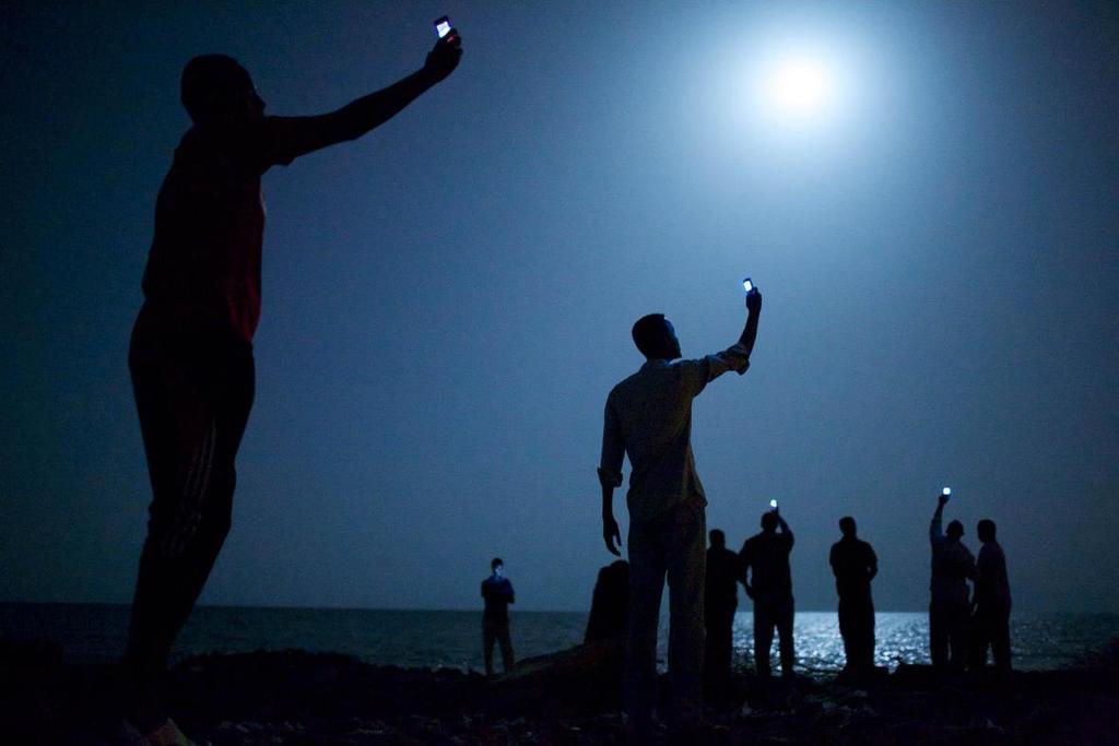 2014 Bildtext: Djibouti City, Djibouti African migrants on the shore of Djibouti City at night raise their phones in an attempt to catch an inexpensive signal from neighboring Somalia a tenuous link