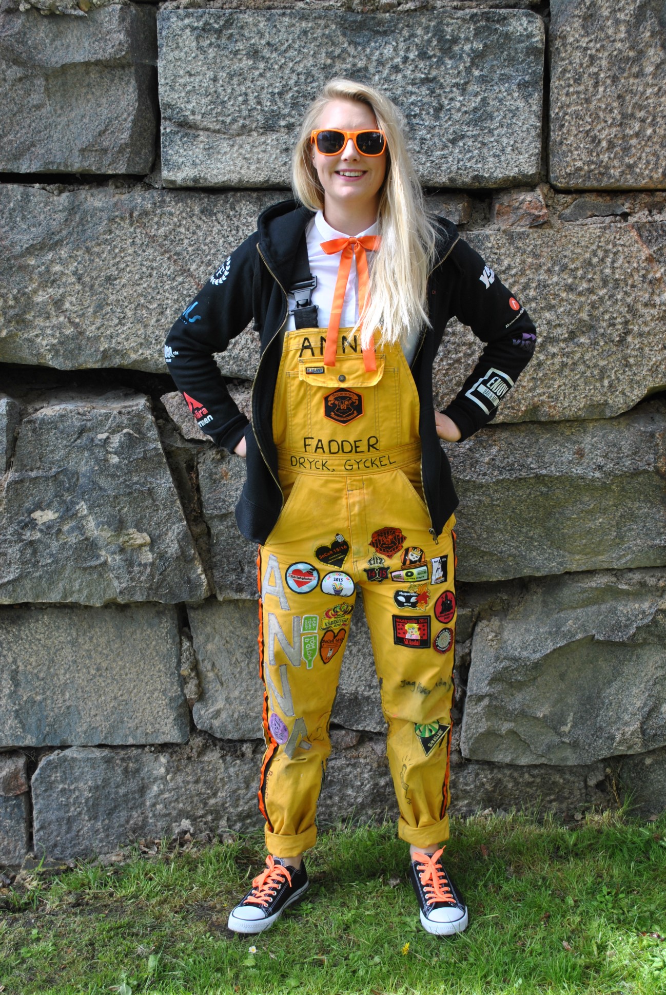 Societeten När solen skiner bär Societeten orangea solglasögon. Societeten går klädd i en svart hoodie som har Societetens eget märke tryckt på ryggen.