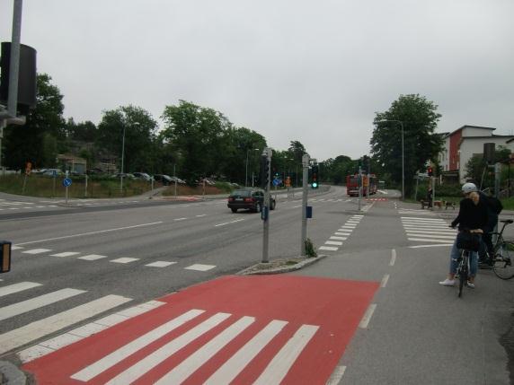 Cyklister som färdas västerut på norra sidan Värmdövägen har nedförsbacke till busshållplats och korsningen.