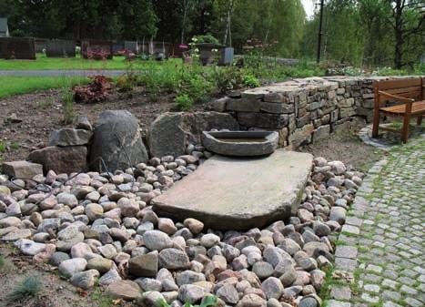 Strax nordost om rabatten och minnesstenen befinner sig en stenlagd yta där en hållare i järnsmide för bland annat buketter är placerad.