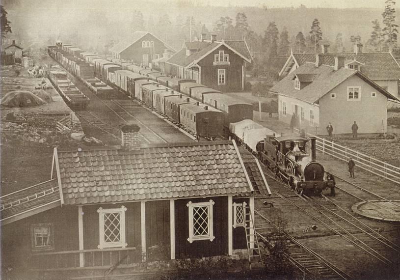 En tidig bild från stationsområdet där två tåg mötas. Vid lastkajen står godsvagnar på båda stickspåren. Bortanför stationshuset ligger godsmagasinet.