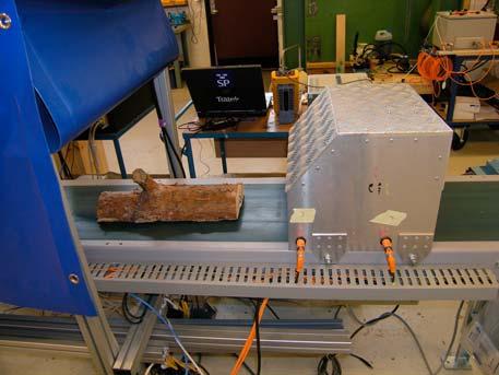 Figur 2.3. Mätuppställningen installerad i SP Träteks laboratorium i Skellefteå. Träbiten ligger på ett transportband och över detta är en huv med mikrovågssensor och fotoceller monterad. Figur 2.