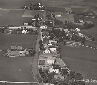 Äldre flygfoto över Alstad längs väg 101, vy mot väster. Alstad skola i bebyggelsegruppen längst bak i bild. Järnvägen illustrerad som streckad linje.