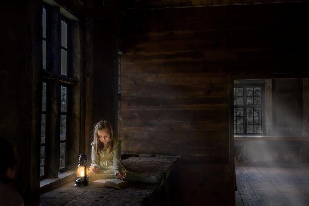 1. Maria Lindberg Upplysning En flicka sitter framför ett fönster på en skrovlig träbänk i ett rum helt i mörkt trä. Framför sig har hon en bok och en lampa.