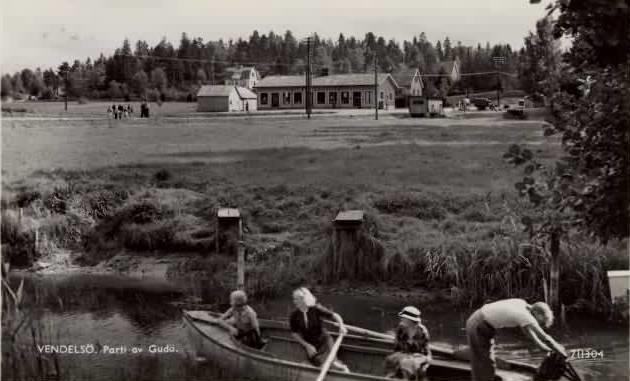 Parti av Gudö Vykortet poststämplat 1957.08.12. I förgrunden Gudöån. Konsum och posten fanns i huset i fonden på 1940-1960-talet. Inrymmer idag Modevillan.