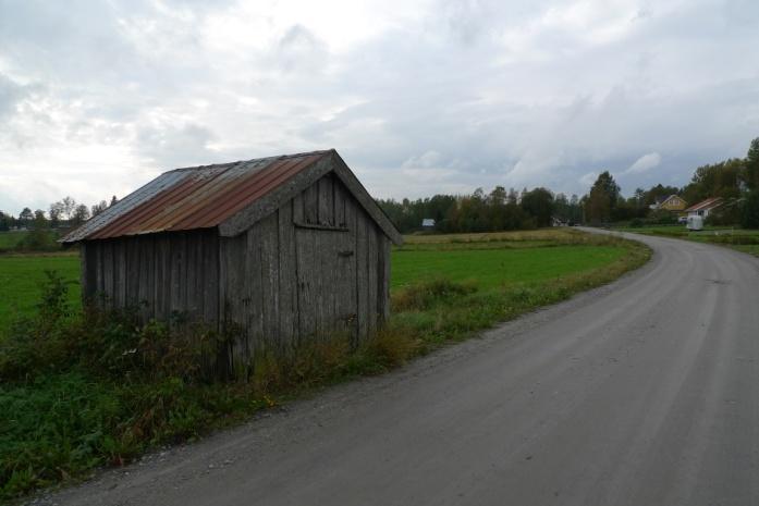 Objekt 52b Brädlada med plåttak som står i kanten mellan en vallåker och en grusväg (foto ovan t.h.).