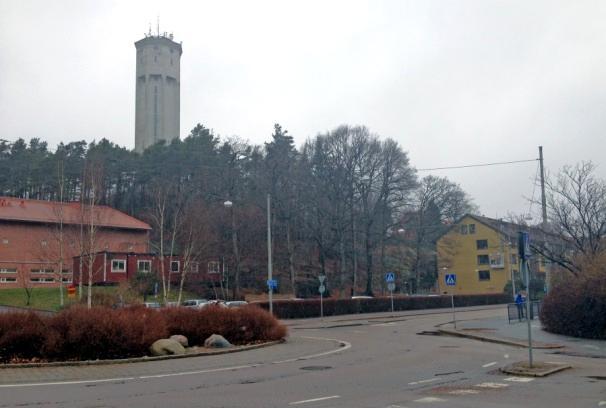 Vy mot tomt intill markparkering som kantas av stora grova träd. Tomten föreslås för ett flerbostadshus med verksamheter. Cirka sju stycken träd kan komma behöva fällas.