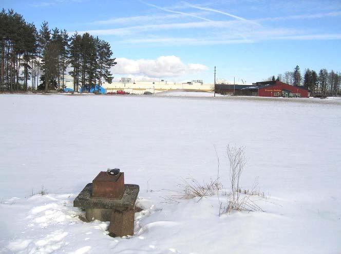 Figur 7. Provtagningspunkt (vid pilen) placerad vid bostäder (dessa syns inte på bilden). I bakgrunden ses fabriksbyggnaden. I fabriken utfördes lackeringsarbeten.