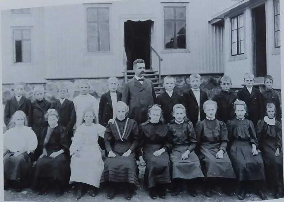 CIRKA 1905 På ett skolfoto några år senare sitter en skolklass framför en ljusmålad träfasad. Vi vet att eleverna är födda 1896. Vi ser endast den nedre delen av byggnaden.